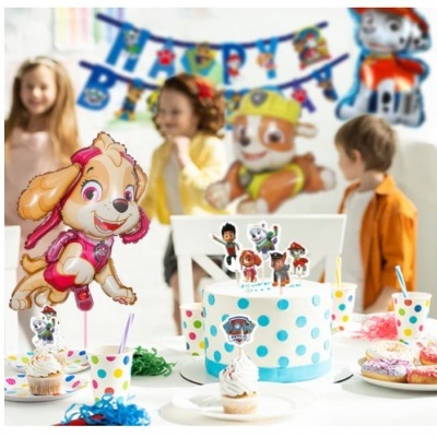 Bolo de aniversário decorado com personagens, copos e cupcakes numa mesa com crianças ao fundo.