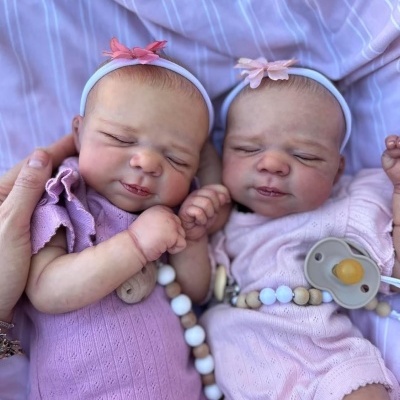 Duas bonecas de bebé com vestidos rosa e lilás e fitas de cabeça com flores cor-de-rosa deitadas sobre um tecido lilás riscado.