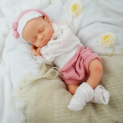 Boneca de bebé vestida de branco e rosa a dormir sobre almofadas e manta branca