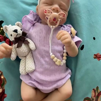 Bebé de brinquedo com roupa lilás e chupeta rosa, segurando urso de croché branco