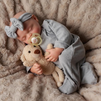 Bebé a dormir vestido com fato cinza e faixa de cabelo, chupeta Mom e segurando ursinho de peluche sobre cobertor bege.