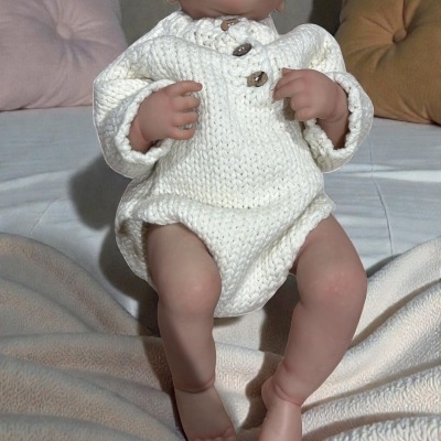 Boneca realista para bebé com roupa de malha branca sentada numa cama.