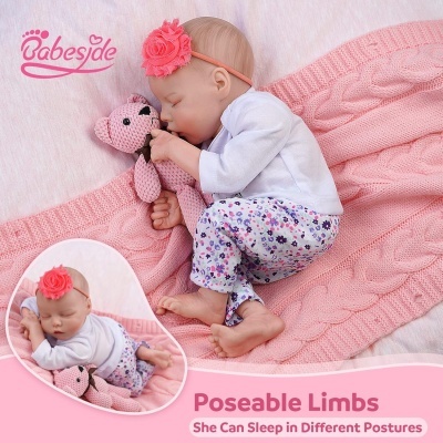 Boneca realista de bebé deitada com roupa branca e leggings florais, segurando ursinho rosa sobre manta rosa trançada