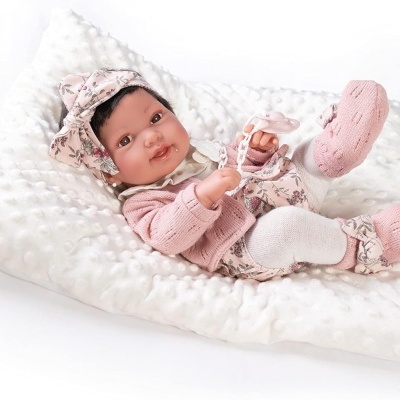 Boneca bebé de roupas cor de rosa e branco deitada numa almofada branca macia.