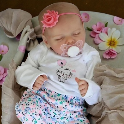 Boneca bebé com roupa branca e rosa, chupeta e fita com flor a dormir sobre tecido e flores