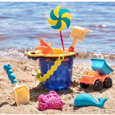 Brinquedos de praia em areia junto ao mar incluindo balde azul, pá, carrinho e brinquedos coloridos.