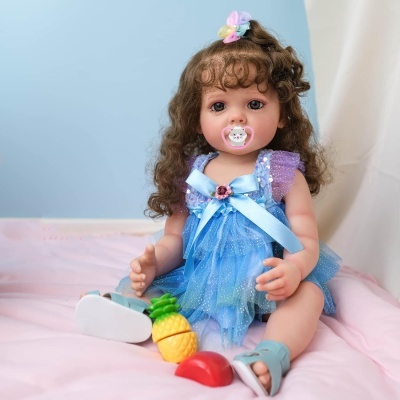 Boneca bebé realista vestida de azul sentada numa cama rosa com brinquedos de frutas à frente