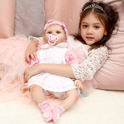 Menina abraçando boneca bebé vestida de branco e rosa, sentadas em almofadas cor-de-rosa.