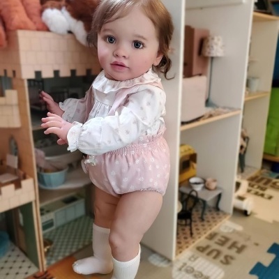 Bebé de brinquedo realista com vestido e calças cor de rosa e brancas junto a casinha de madeira