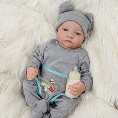 Boneco bebé com pijama cinzento e biberão branco deitado em superfície felpuda branca