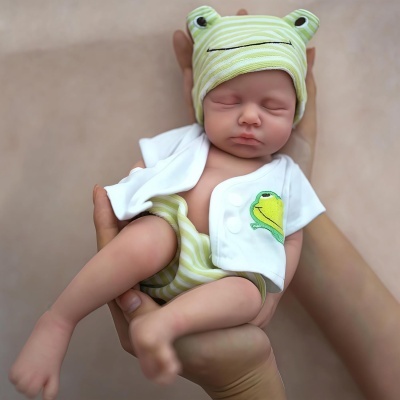 Boneca bebé realista com chapéu verde riscado, fralda às riscas e camisola branca com bordado de sapo