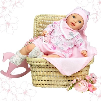 Boneca bebê realista com pijama rosa sentada numa cesta de palha com manta rosa ao lado de um cavalo de balanço cor-de-rosa e flores