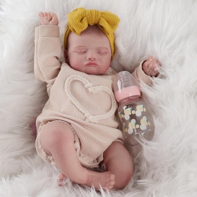 Boneca bebé realista com camisola bege e faixa amarela, deitada em pelúcia e segurando mamadeira rosa