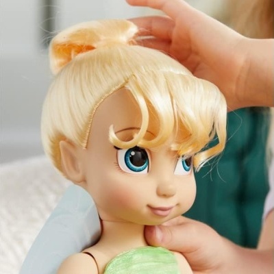 Boneca com cabelo loiro, olhar azul e vestido verde sendo ajustada por mãos adultas