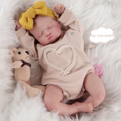 Boneca bebé vestindo body bege com coração, fita amarela, e ursinho de malha ao lado numa superfície branca felpuda.