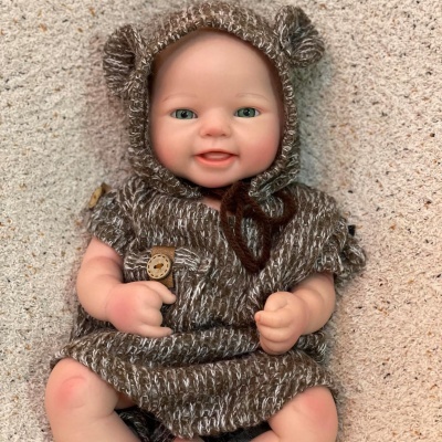 Boneca bebé com fato de malha castanho e bege com capuz e botão, deitada sobre tecido macio bege.