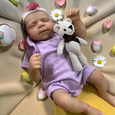 Boneca bebé realista com roupa lilás e ursinho cinzento sobre manta bege com flores