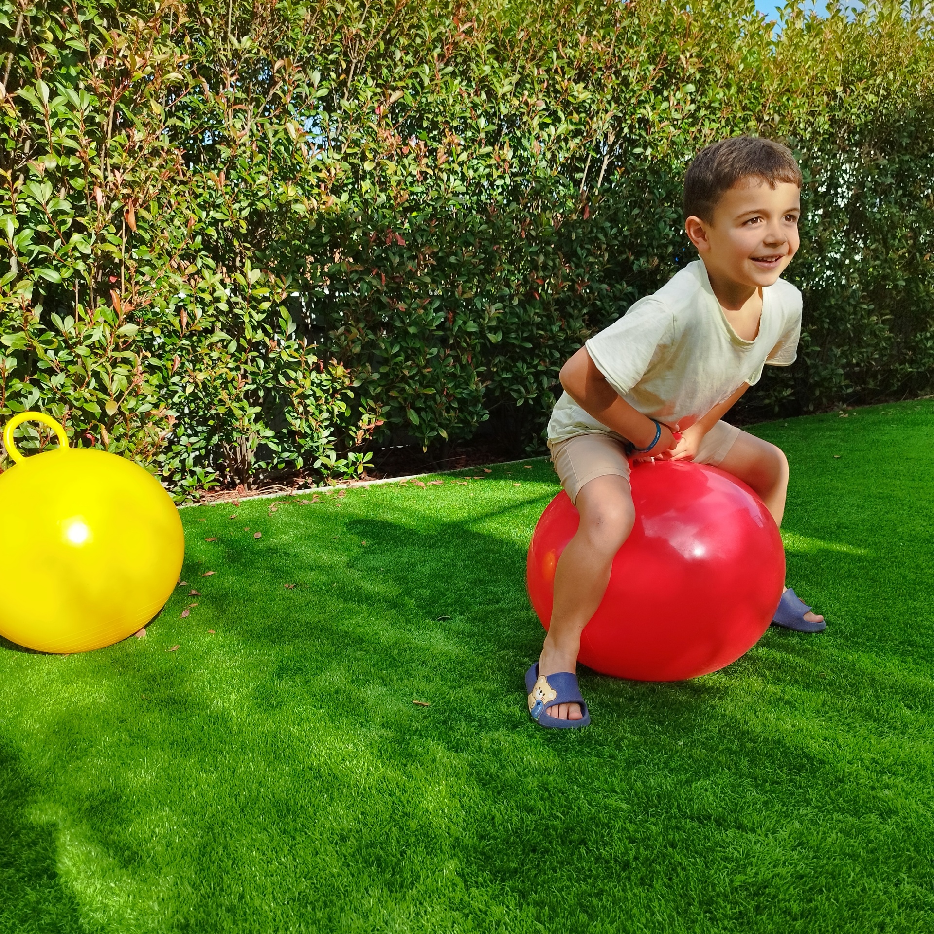 BOLA CANGURU SALTITONA 45CM, SOLMAR Criança em balão de salto vermelho em relvado verde com balão amarelo ao lado e arbustos ao fundo