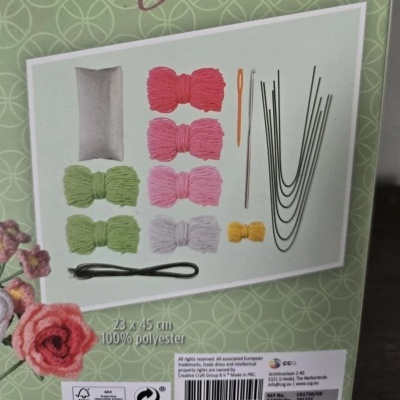 Kit de materiais com fios coloridos, agulha, arame e saco branco em embalagem com informações de 23 x 45 cm e 100% poliéster.