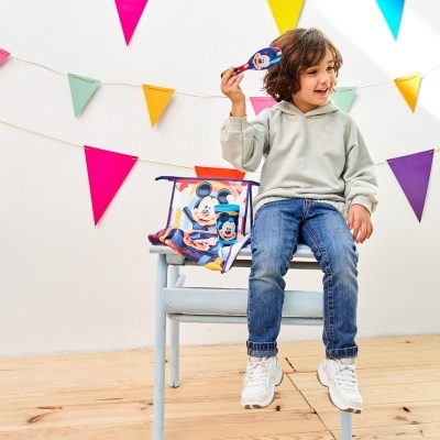 Criança com máscara e mochila do Mickey Mouse sentada numa cadeira azul com bandeirolas coloridas no fundo