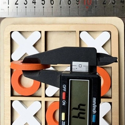 Jogo tic-tac-toe em madeira com peças brancas e laranja e paquímetro digital a medir peça.