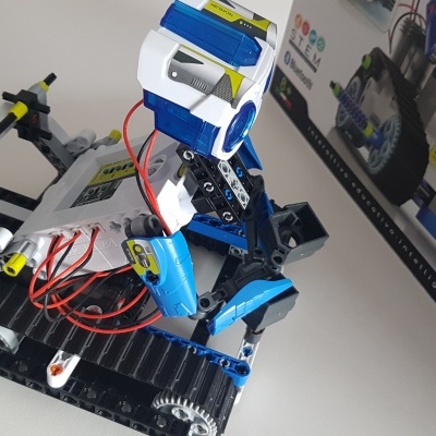 Robô de plástico azul, branco e preto com fios vermelhos e base com lagartas, junto a caixa do produto.