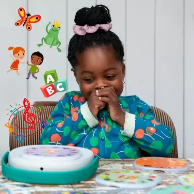 Criança brincando com placas coloridas numa mesa, vestindo casaco azul com cerejas e cabelo preso com fita rosa.