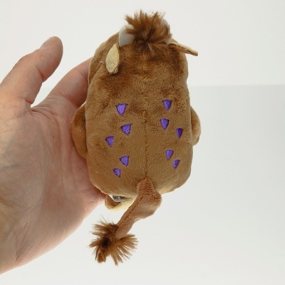 Pelúcia castanha de animal com manchas roxas e cauda, segurada por uma mão