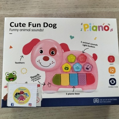 Brinquedo musical infantil em forma de cão rosa com botões coloridos, numa caixa branca com texto e ícones
