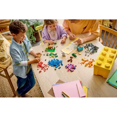 Crianças e adulto a brincar com peças LEGO coloridas numa mesa de madeira com estojo amarelo LEGO e material de desenho.