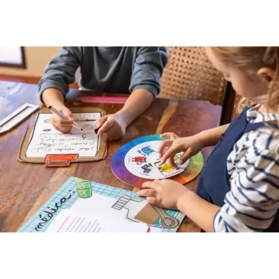 Crianças a usar materiais educativos em mesa de madeira, com disco colorido de emoções e prancha de desenho com texto em português.