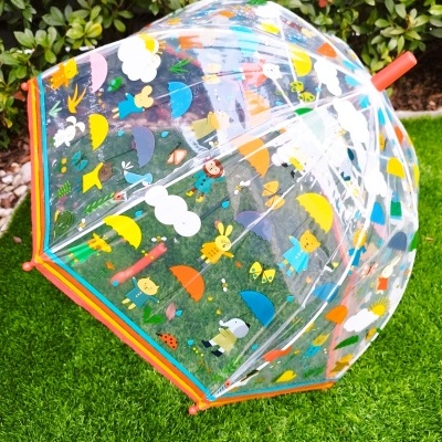Guarda-chuva infantil transparente com estampas coloridas e bordas laranja e azul sobre relva.