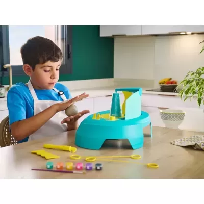 Criança a trabalhar massa cinzenta com aparelho azul e ferramentas coloridas numa cozinha moderna.