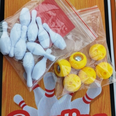 Mini pinos de bowling brancos e bolas amarelas com rolamento, sobre tapete decorativo madeira com a palavra Bowling.