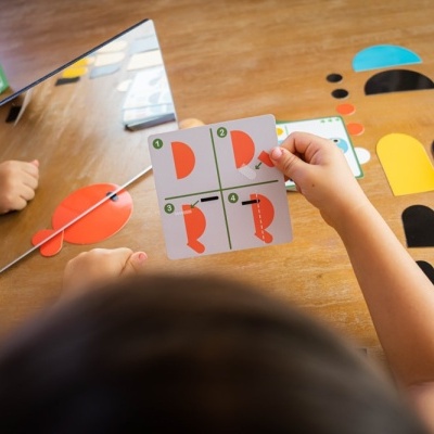Jogo infantil com cartas, espelho e peças coloridas sobre mesa de madeira.