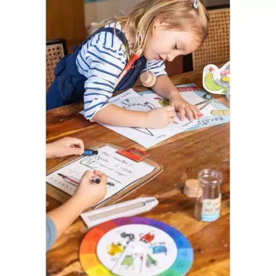 Criança a desenhar enquanto outra mão escreve numa lista sobre mesa com discos coloridos e garrafa de vidro