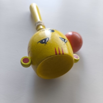 Maraca de madeira amarela com olhos desenhados e bola laranja na lateral