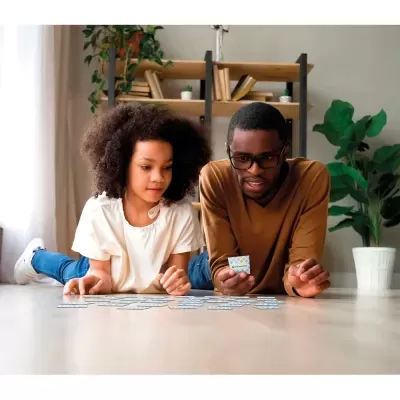 Duas pessoas a jogar cartas com padrão azul e branco no chão, rodeadas por estante, livros e plantas.