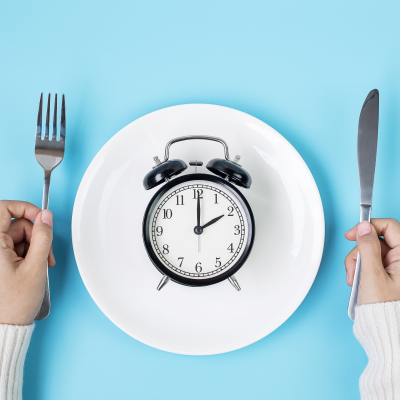 Relógio despertador num prato branco com fundo azul e talheres nas mãos