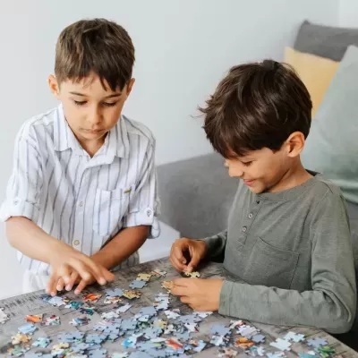 Dois miúdos montam um puzzle numa mesa cinzenta, com um sofá e almofadas ao fundo.