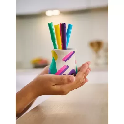 Recipiente cerâmico com decoração colorida e canetas coloridas dentro, segurado por mãos.
