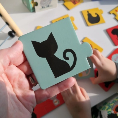 Peça de puzzle verde com desenho preto de gato segurada por uma mão próximo das mãos de uma criança