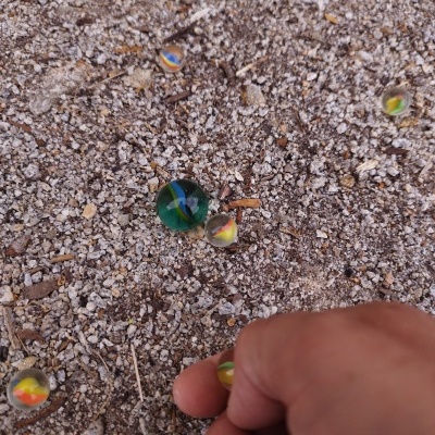 Berlindes de vidro coloridos sobre chão de terra e pedras, mão segura um berlinde