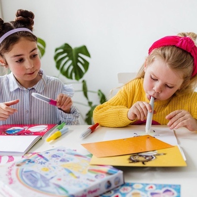 Duas crianças a desenhar e pintar numa mesa branca com marcadores coloridos