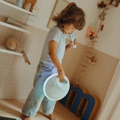 Menina em cima de móvel segurando brinquedo redondo azul claro e boneca, roupa às riscas, móvel com guitarra azul e letra m azul.