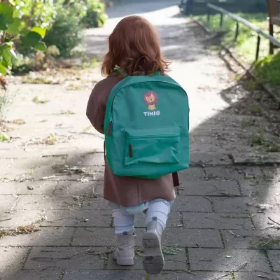 Mochila infantil verde com bolso frontal e desenho de leão, usada por criança vestida com casaco castanho e botas cinzentas