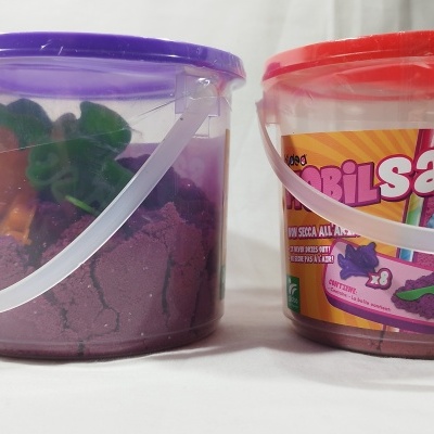 Dois baldes de areia colorida para brincadeira, um com tampa roxa e outro vermelha, com etiquetas visíveis.