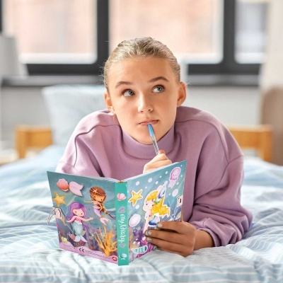 Caderno infantil com capa azul e ilustrações de sereias e peixes, menina com casaco lilás a segurar o caderno deitada numa cama