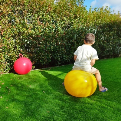 Criança sentada em bola de saltar amarela num relvado com bola vermelha ao lado e sebe ao fundo
