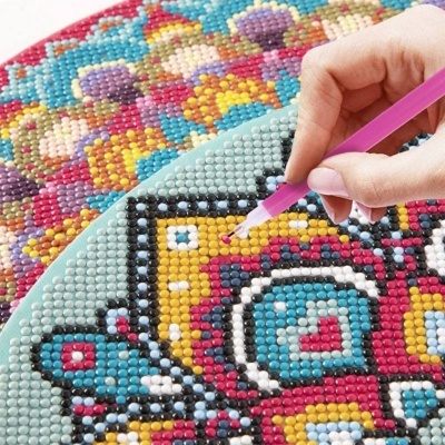 Mãos aplicando pequenos cristais coloridos numa tela com padrão geométrico e floral